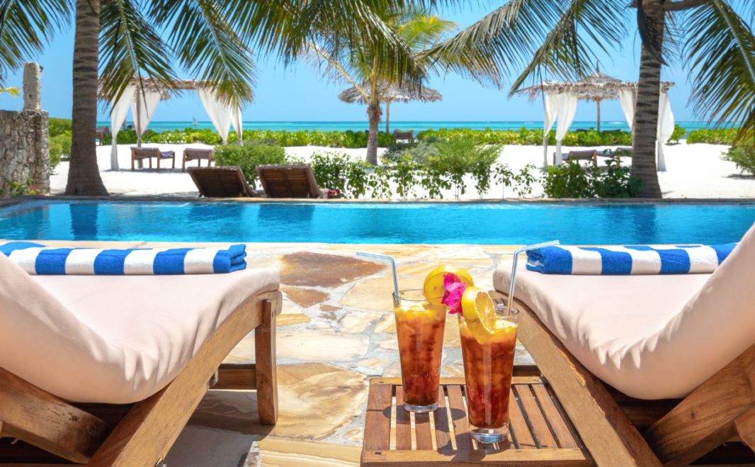 Image of two sun loungers facing a swimming pool with a tropical scene in the background. Two cocktails stand on a table beside the sun loungers.