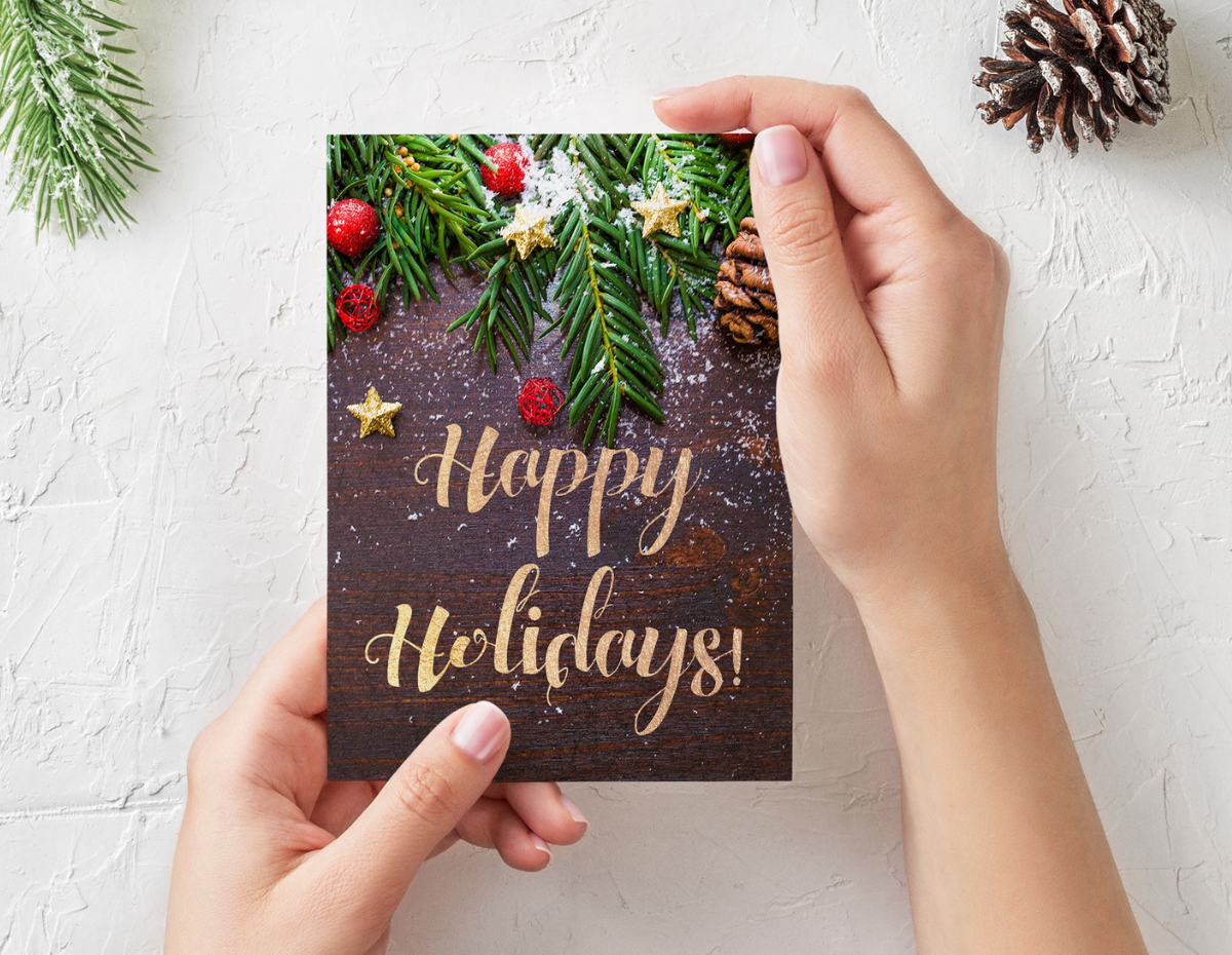 Image of two hands holding a Christmas card with the words Happy Holidays and festive foliage.