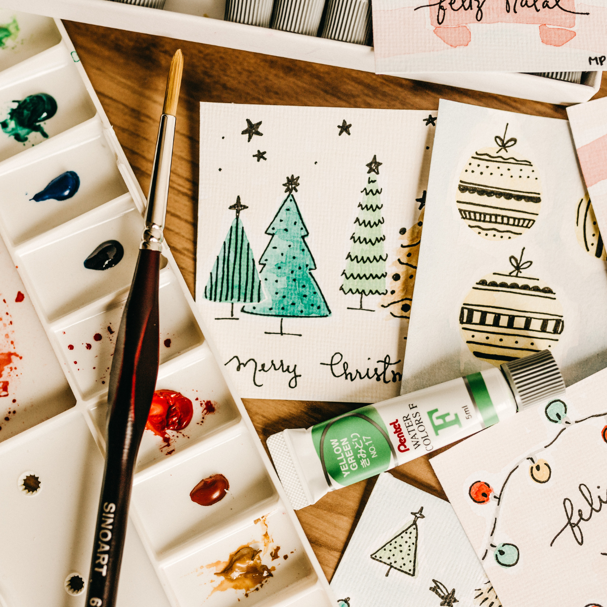 A selection of Christmas cards with various hand painted designs, a paintbrush and paint palette lies alongside the cards.